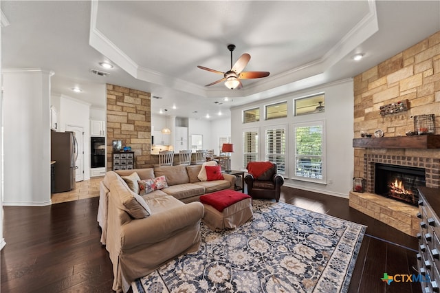 living room with ceiling fan, a stone fireplace, dark hardwood / wood-style flooring, crown molding, and a tray ceiling