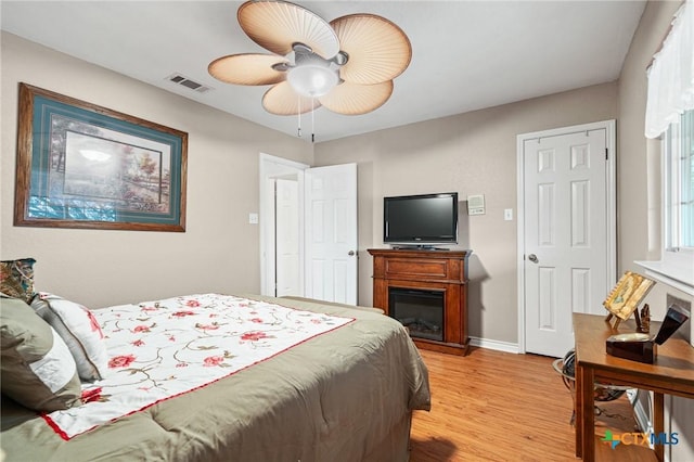 bedroom featuring light wood-type flooring, a glass covered fireplace, baseboards, visible vents, and a ceiling fan
