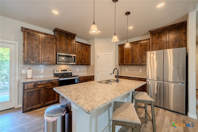 kitchen featuring an island with sink, appliances with stainless steel finishes, light stone counters, decorative light fixtures, and a sink