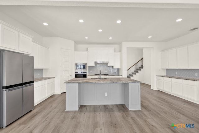 kitchen featuring stainless steel appliances, white cabinets, decorative backsplash, an island with sink, and light wood-type flooring