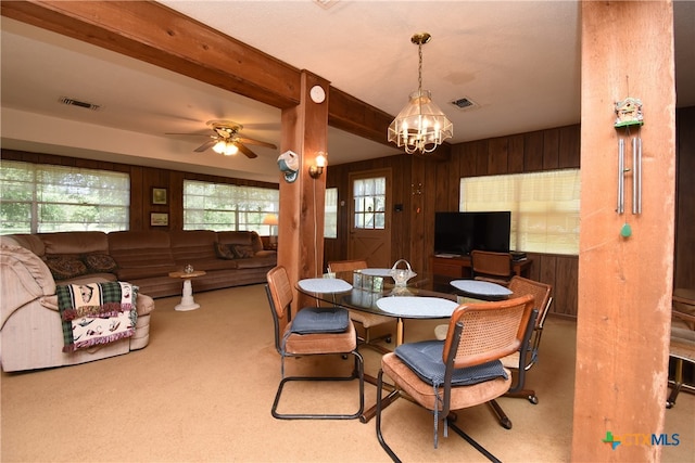 dining area with ceiling fan with notable chandelier, beamed ceiling, light carpet, and wooden walls