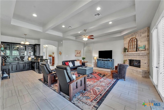 living room with a stone fireplace, beamed ceiling, indoor bar, and ceiling fan with notable chandelier