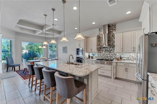 kitchen with light stone countertops, hanging light fixtures, wall chimney range hood, a kitchen island with sink, and appliances with stainless steel finishes