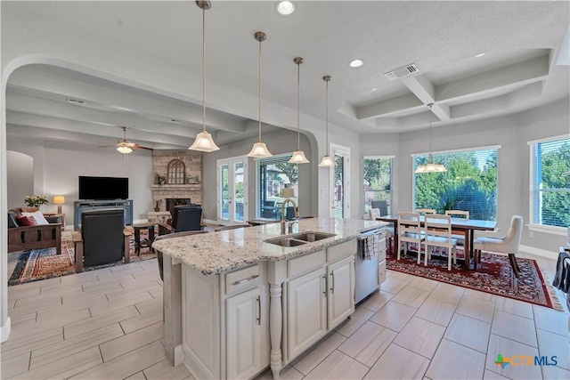 kitchen featuring ceiling fan, sink, a center island with sink, a stone fireplace, and hanging light fixtures