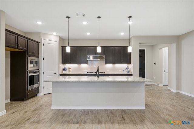 kitchen with stainless steel appliances, light stone countertops, dark brown cabinets, and a center island with sink