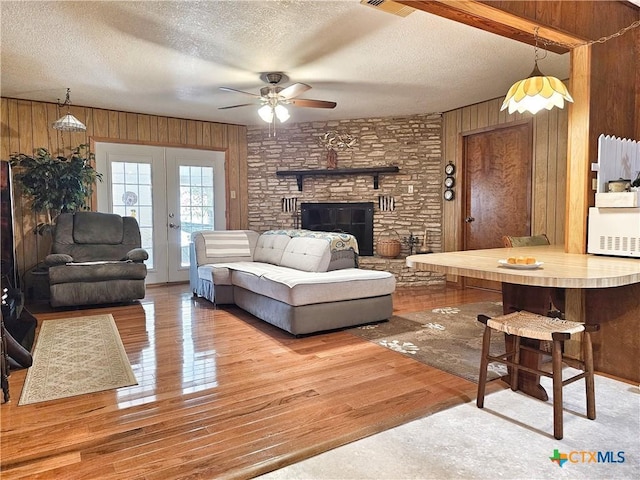 living room featuring ceiling fan, a stone fireplace, wood-type flooring, a textured ceiling, and wooden walls