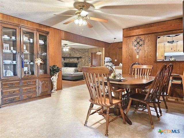 dining room with ceiling fan, wood walls, a stone fireplace, and a textured ceiling