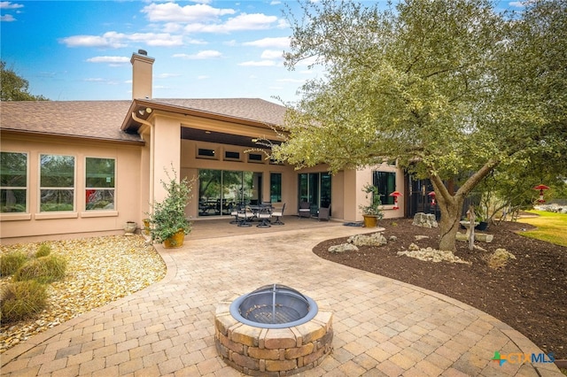 back of property with a patio, a fire pit, a shingled roof, stucco siding, and a chimney