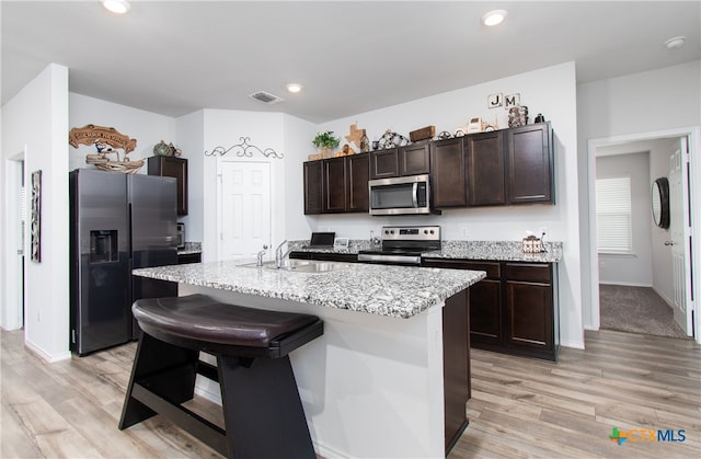 kitchen with dark brown cabinetry, appliances with stainless steel finishes, a kitchen bar, an island with sink, and light wood-type flooring