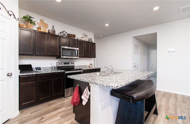 kitchen with light wood-type flooring, appliances with stainless steel finishes, dark brown cabinetry, sink, and a kitchen island with sink