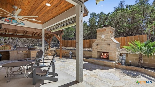 view of patio featuring grilling area, fence, exterior kitchen, an outdoor stone fireplace, and a ceiling fan