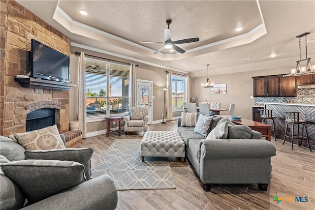 living room with ceiling fan with notable chandelier, a raised ceiling, crown molding, a fireplace, and light hardwood / wood-style floors