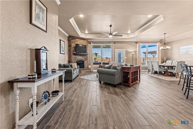 living room with ceiling fan with notable chandelier, a stone fireplace, a raised ceiling, crown molding, and plenty of natural light