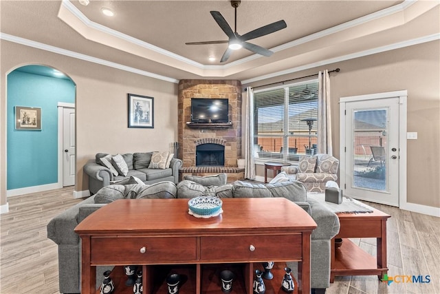 living room featuring a stone fireplace, ceiling fan, ornamental molding, a tray ceiling, and light hardwood / wood-style floors