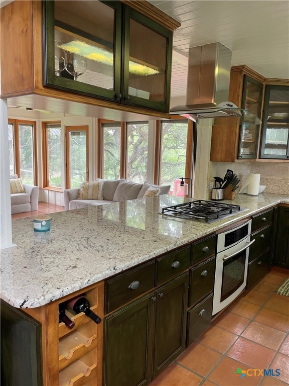 kitchen featuring island range hood, light tile patterned flooring, backsplash, appliances with stainless steel finishes, and light stone countertops