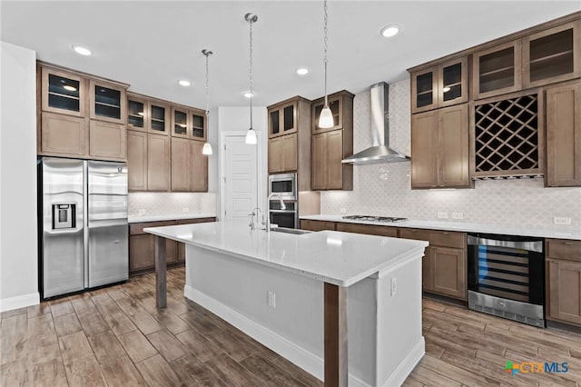 kitchen with beverage cooler, pendant lighting, stainless steel appliances, wall chimney exhaust hood, and an island with sink