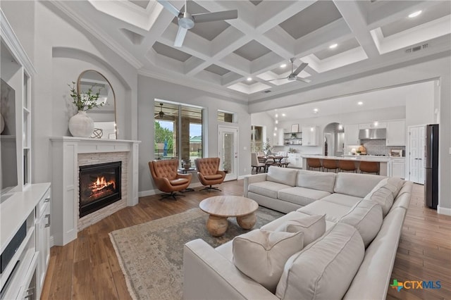 living area featuring light wood-style floors, a stone fireplace, a ceiling fan, a towering ceiling, and beamed ceiling