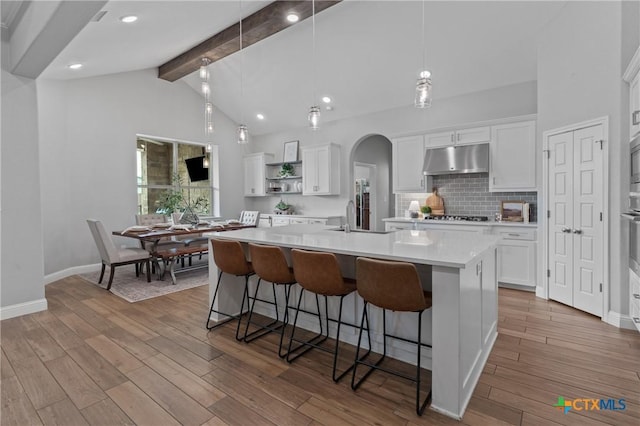 kitchen with open shelves, a center island with sink, beam ceiling, light countertops, and under cabinet range hood