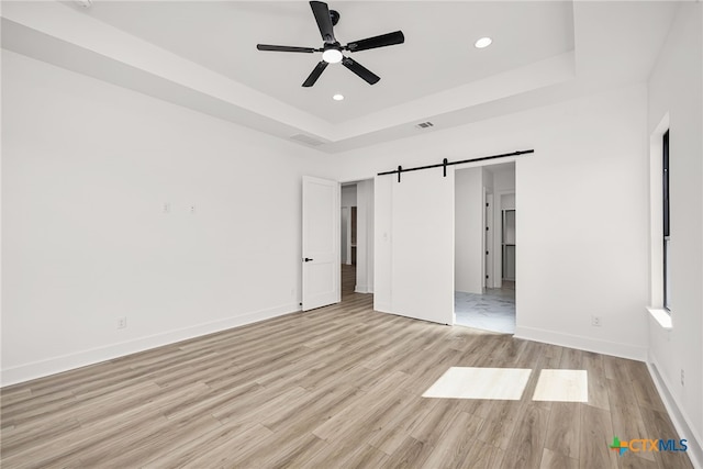 unfurnished bedroom featuring light hardwood / wood-style flooring, a raised ceiling, a barn door, and ceiling fan