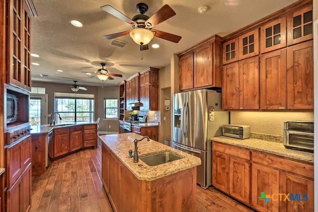 kitchen with sink, dark hardwood / wood-style floors, light stone countertops, a center island with sink, and appliances with stainless steel finishes