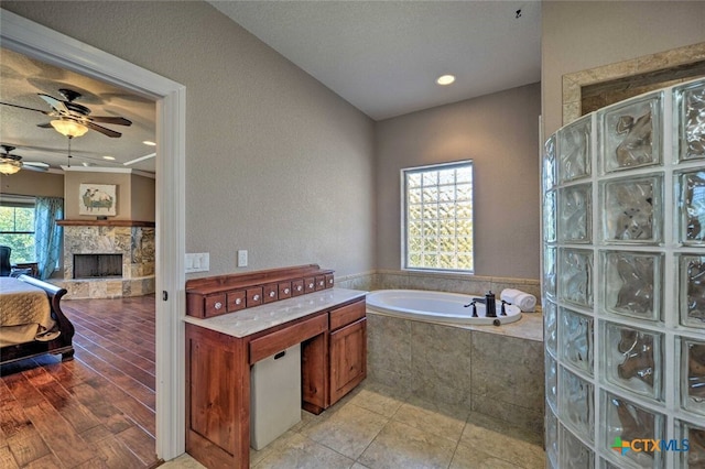 bathroom with a relaxing tiled tub, ceiling fan, and a stone fireplace