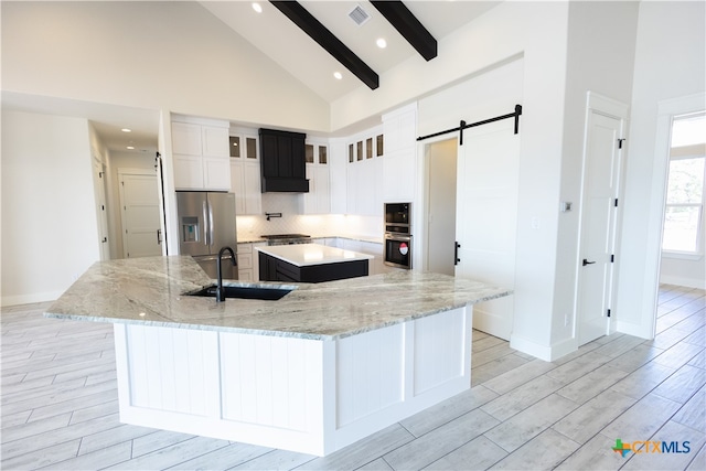 kitchen with light stone countertops, stainless steel refrigerator with ice dispenser, a large island with sink, a barn door, and beam ceiling