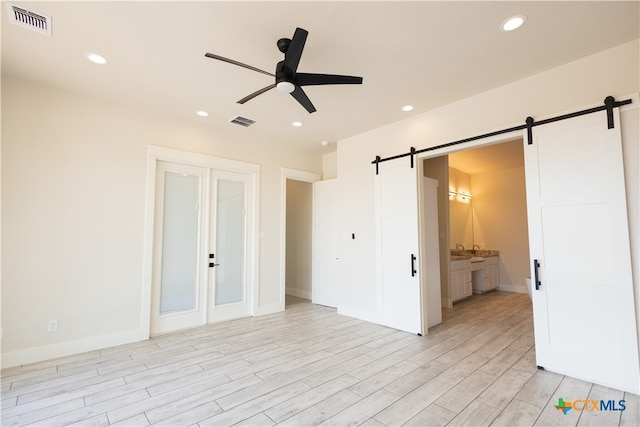 unfurnished bedroom featuring a barn door, light hardwood / wood-style flooring, ensuite bath, and ceiling fan