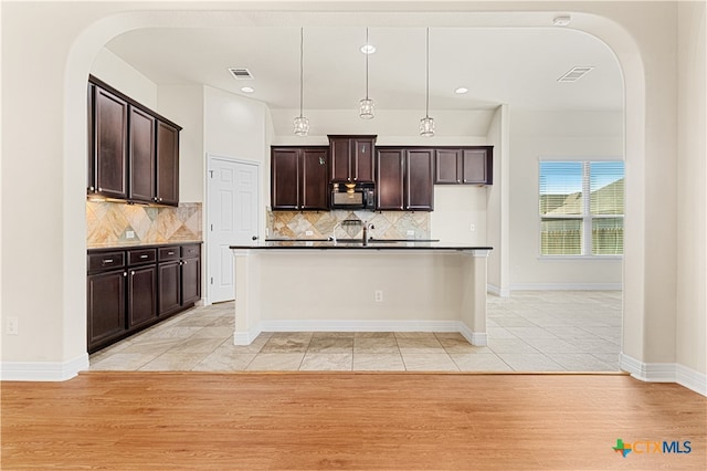 kitchen with a center island with sink, hanging light fixtures, tasteful backsplash, dark brown cabinets, and light hardwood / wood-style floors