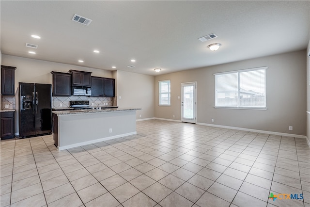 kitchen with black appliances, a center island with sink, light tile patterned floors, light stone countertops, and decorative backsplash