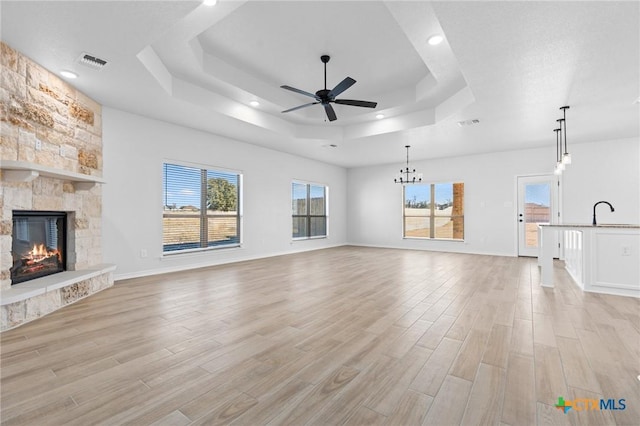 unfurnished living room with ceiling fan, a stone fireplace, a raised ceiling, and light hardwood / wood-style flooring