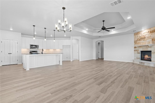 unfurnished living room featuring a stone fireplace, sink, light wood-type flooring, a tray ceiling, and ceiling fan with notable chandelier