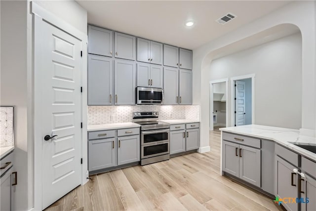 kitchen featuring gray cabinets, appliances with stainless steel finishes, tasteful backsplash, light stone countertops, and light hardwood / wood-style flooring