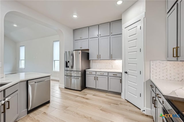 kitchen featuring appliances with stainless steel finishes, gray cabinetry, backsplash, light stone countertops, and light hardwood / wood-style flooring