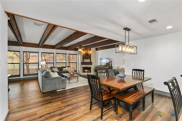 dining space with ceiling fan, a stone fireplace, wood finished floors, and beam ceiling