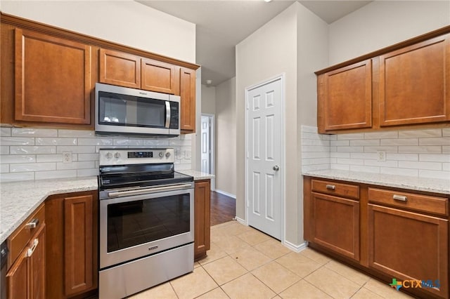 kitchen with brown cabinetry, light stone counters, stainless steel appliances, and backsplash