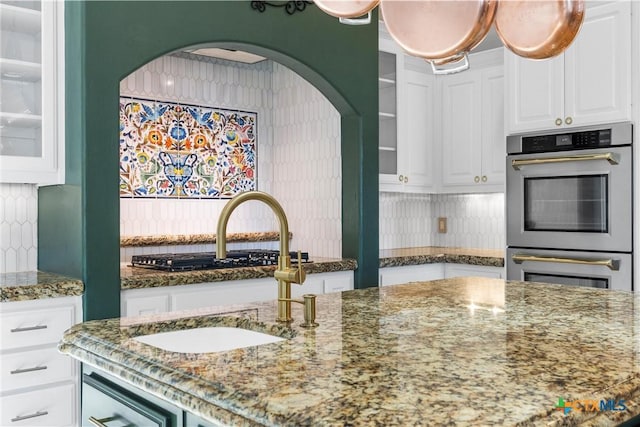 kitchen with dark stone counters, white cabinets, glass insert cabinets, double oven, and a sink