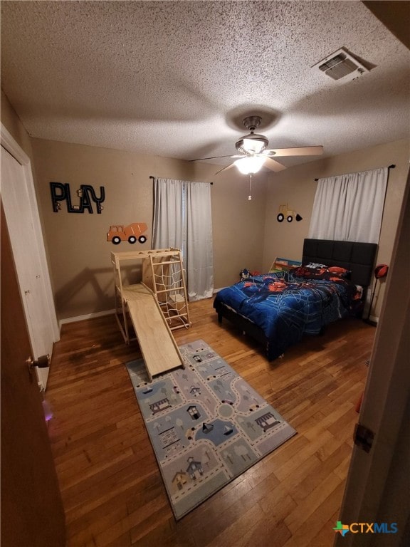 bedroom with ceiling fan, a textured ceiling, visible vents, wood finished floors, and baseboards