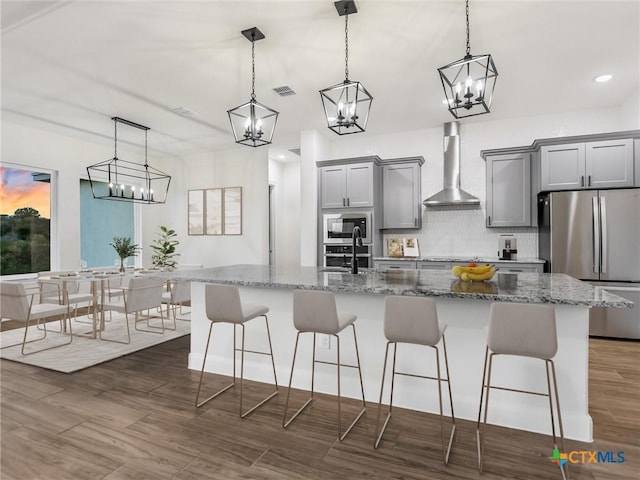 kitchen featuring stainless steel appliances, wall chimney exhaust hood, a center island with sink, and decorative light fixtures