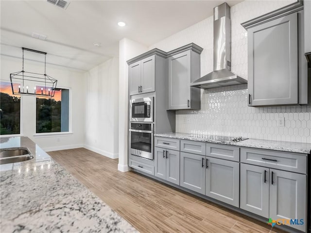 kitchen featuring stainless steel appliances, light stone countertops, pendant lighting, light hardwood / wood-style flooring, and wall chimney range hood