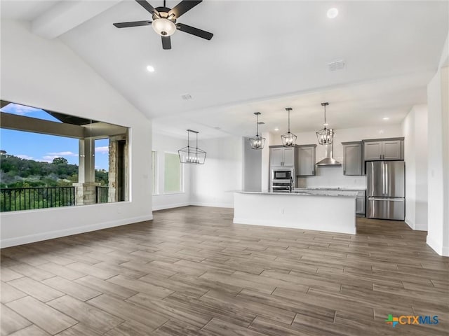 kitchen with appliances with stainless steel finishes, wall chimney exhaust hood, hanging light fixtures, a kitchen island with sink, and gray cabinetry