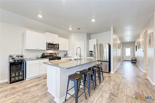 kitchen with stainless steel appliances, a center island with sink, beverage cooler, white cabinets, and light stone countertops