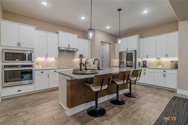 kitchen with a center island with sink, white cabinets, appliances with stainless steel finishes, decorative light fixtures, and light stone counters