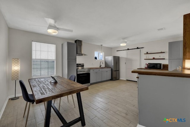 kitchen featuring a barn door, appliances with stainless steel finishes, gray cabinetry, wall chimney range hood, and a sink