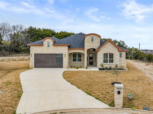 french provincial home featuring roof with shingles, concrete driveway, a garage, stone siding, and a front lawn