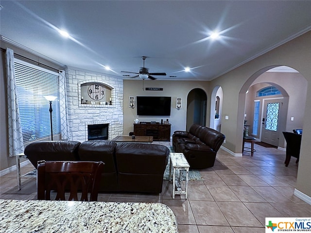 living room with a stone fireplace, tile patterned flooring, ceiling fan, and crown molding