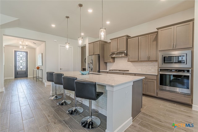 kitchen with appliances with stainless steel finishes, hanging light fixtures, a center island with sink, light stone countertops, and a kitchen bar