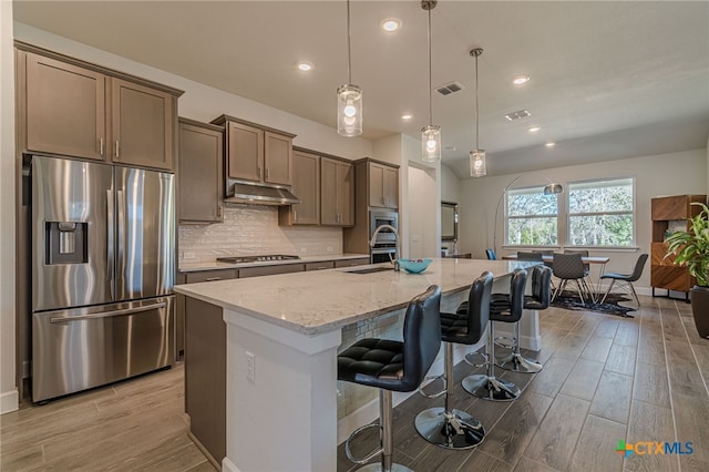 kitchen with a kitchen bar, appliances with stainless steel finishes, pendant lighting, light stone countertops, and a kitchen island with sink