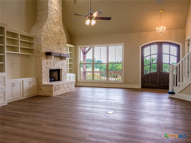 unfurnished living room featuring a stone fireplace, ceiling fan with notable chandelier, dark wood-type flooring, high vaulted ceiling, and built in features