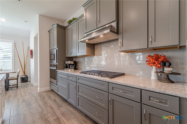 kitchen featuring backsplash, light hardwood / wood-style floors, appliances with stainless steel finishes, gray cabinetry, and light stone counters