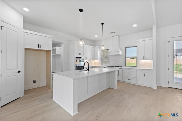 kitchen featuring premium range hood, white cabinetry, light wood-type flooring, pendant lighting, and an island with sink